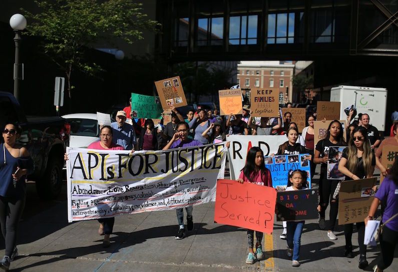 An image of 70 community members gather together, there is a banner at the front that says "APIs for Justice"