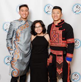 Three people pose at a gala, the person on the left and right are wearing traditional clothing and the person in the middle is wearing a black dress