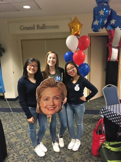 Women smiling while holding a head poster of Hillary Clinton.