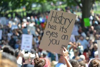 Hand holds a sign stating History has its eyes on you