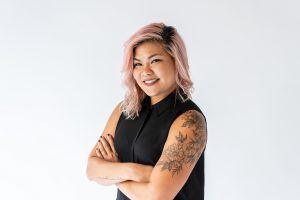 A photo of community leader Tracy Nguyen. Tracy has pink, shoulder length hair and is wearing a black sleeveless blouse. Her arms are crossed and she is smiling at the camera.