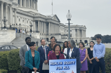 An individual stands in front a podium that says Health Equity for All. A group of individuals stand behind. The Capitol Building is in the background.