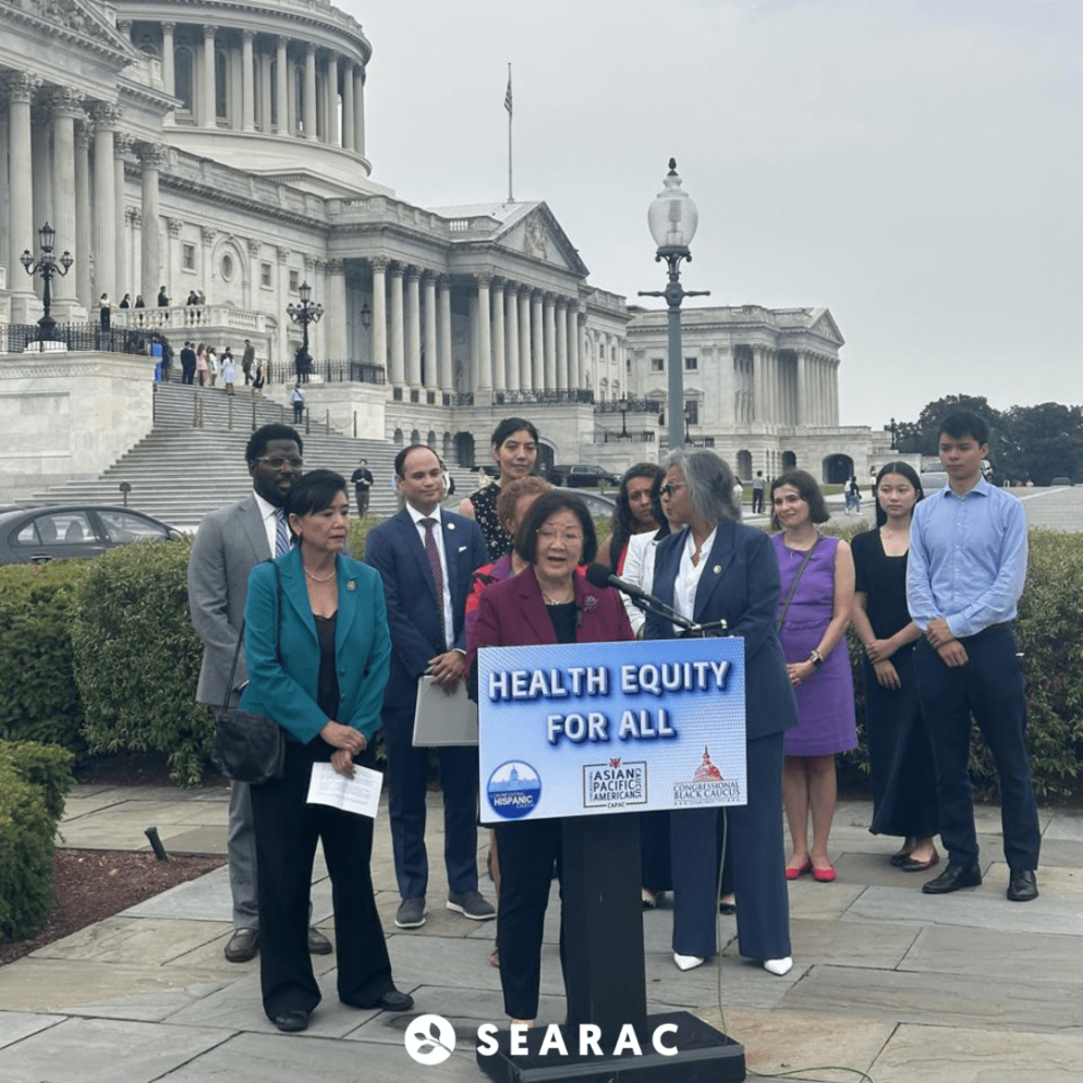 An individual stands in front a podium that says Health Equity for All. A group of individuals stand behind. The Capitol Building is in the background.