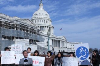 Person speaks at podium in front of sign-holding people at the Capitol