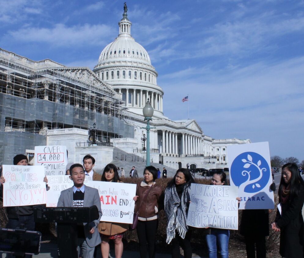 Person speaks at podium in front of sign-holding people at the Capitol