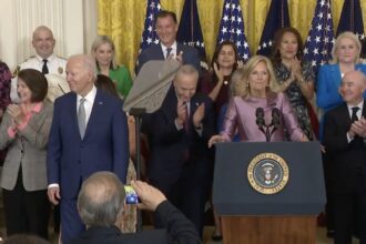 A group of individuals surround President Biden in applause.