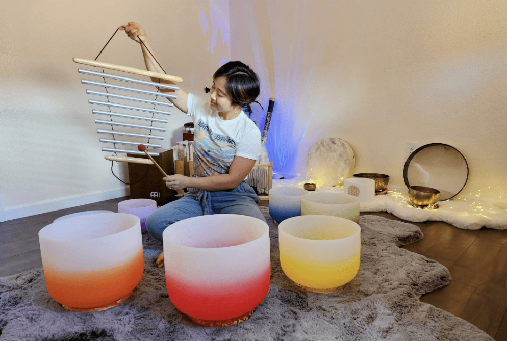 An individual holds up an instrument while sitting behind a circle of brightly colored singing bowls