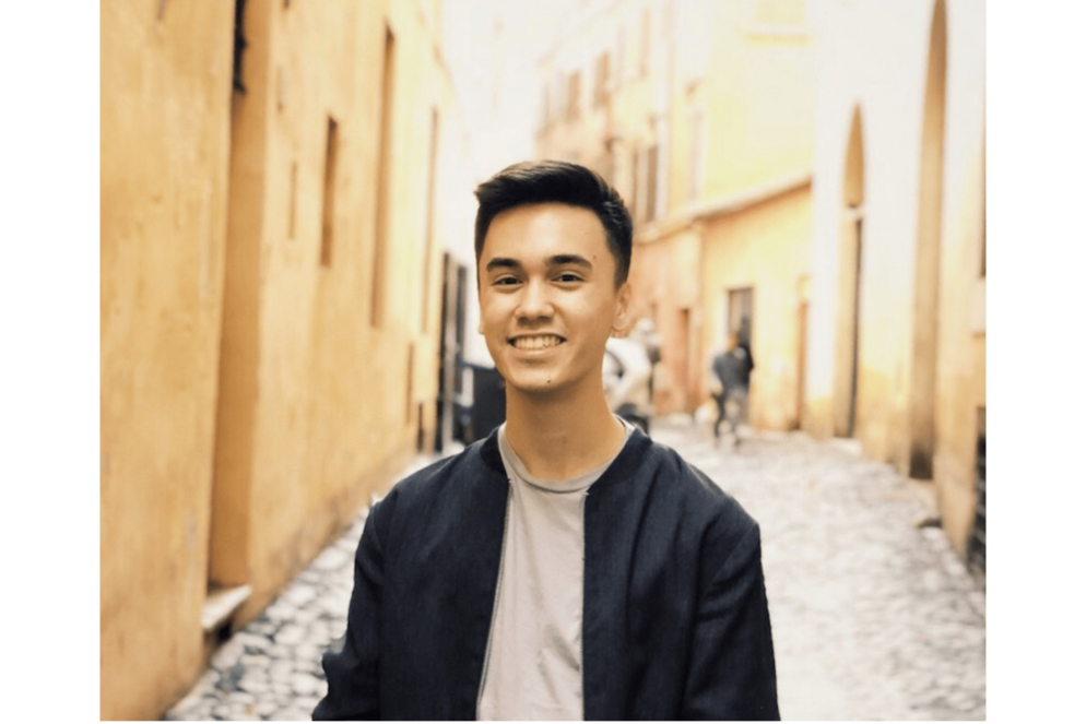 A person wearing a black jacket poses in an alley of yellow colored buildings.