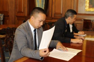 A person in a gray suit and tie holds a document and looks at a document on a desk. Another individual sits adjacently.