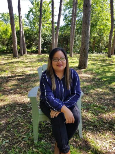 Person with striped blue and white shirt and black pants sits on a chair amongst trees