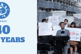 Person speaks at podium, crowd behind holds signs against deportation