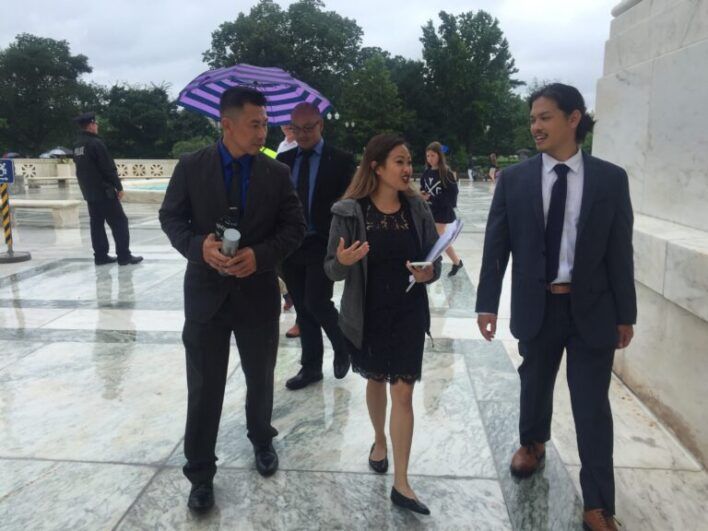 Ke dressed in a suit walks with others, umbrella in the background