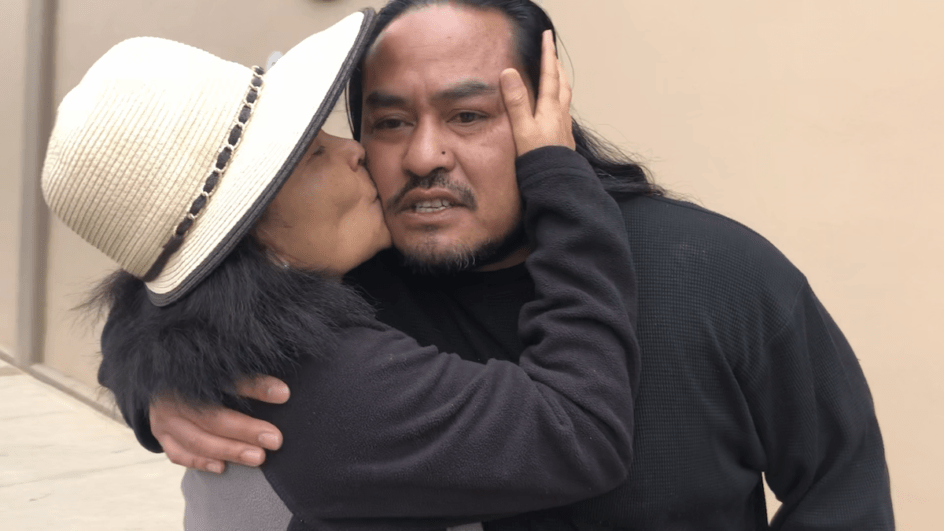 A mother wearing a hat tenderly kisses her son, wearing a black tshirt, on the cheek 