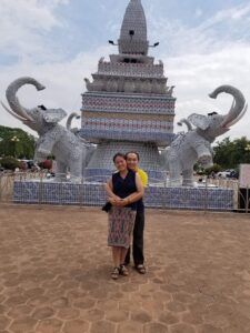 Moua and Dokmai embracing in front of a monument with two white elephant statues.