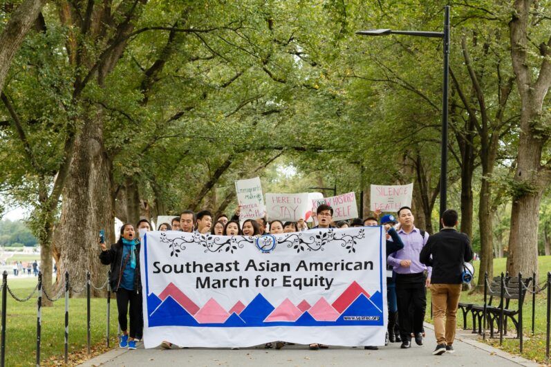 People march through a tree-lined road, holding Southeast Asian American March for Equity sign