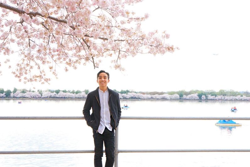 An image of a person dressed in a black jacket, black pants, and a white shirt with cherry blossoms in bloom and a river behind them 