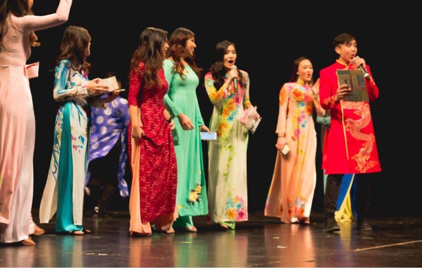 A group of young people wearing traditional Vietnamese outfits and a person at the front a red outfit with a mic