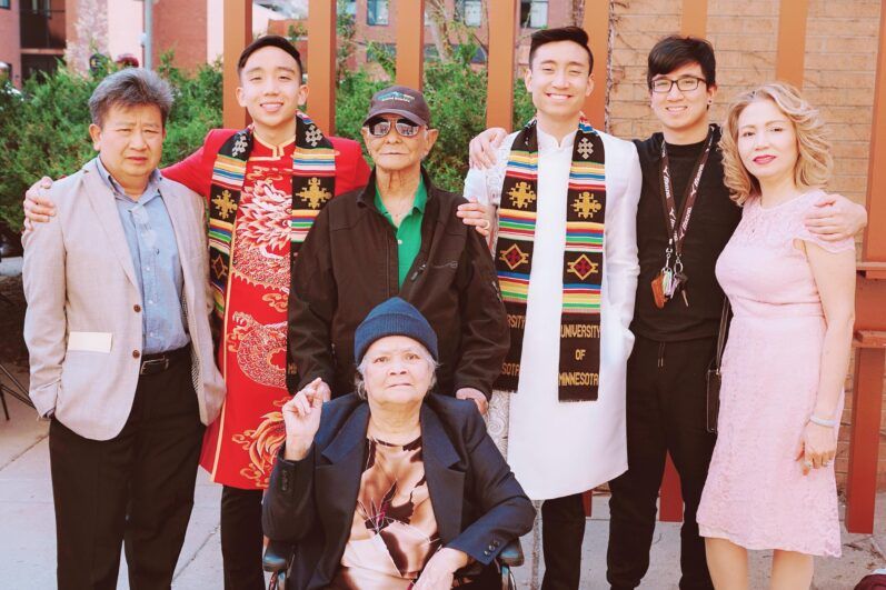 A family smiles together during a graduation ceremony, the graduates wear traditional clothing their graduation stoles