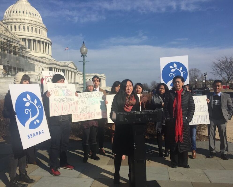 A leader speaking at a podium with people holding signs in the back