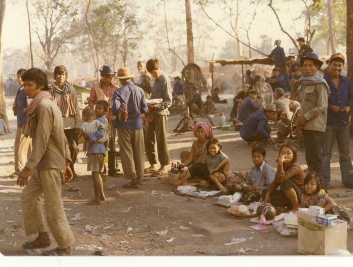 People sit on the ground and stand in a refugee camp