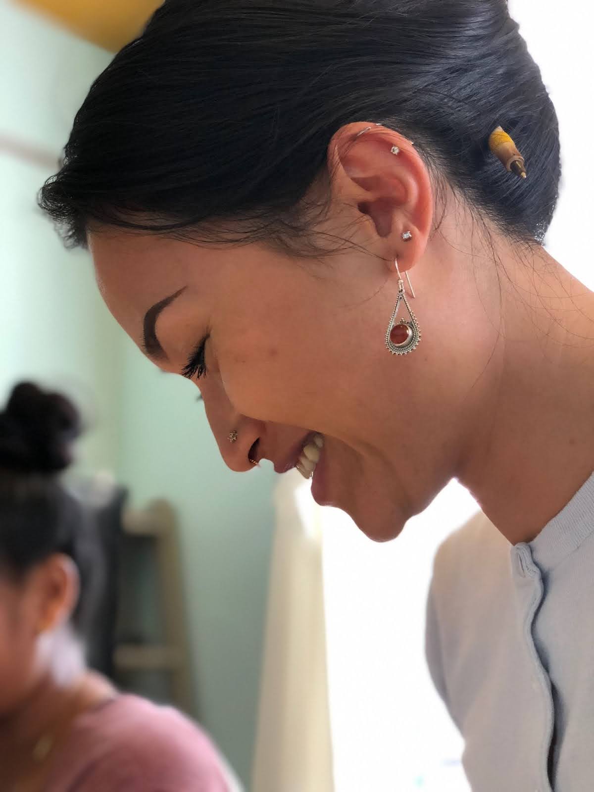 A side profile of a young woman smiling. She is wearing silver earrings with a red pendant and her hair is tied up with a pen. 