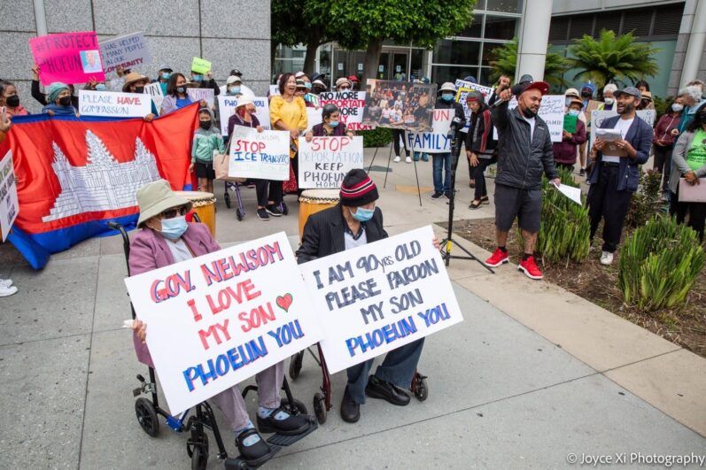 A demonstration with two elders at the front holding signs for the governor to pardon their child. 