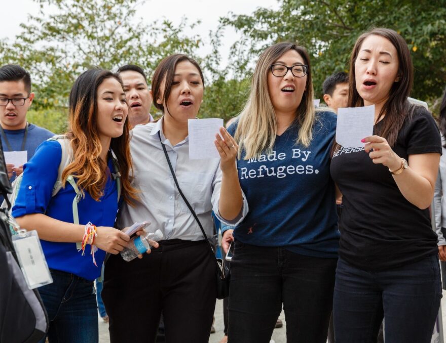 four individuals read from a script, one individual wearing a t-shirt that says Made by Refugees