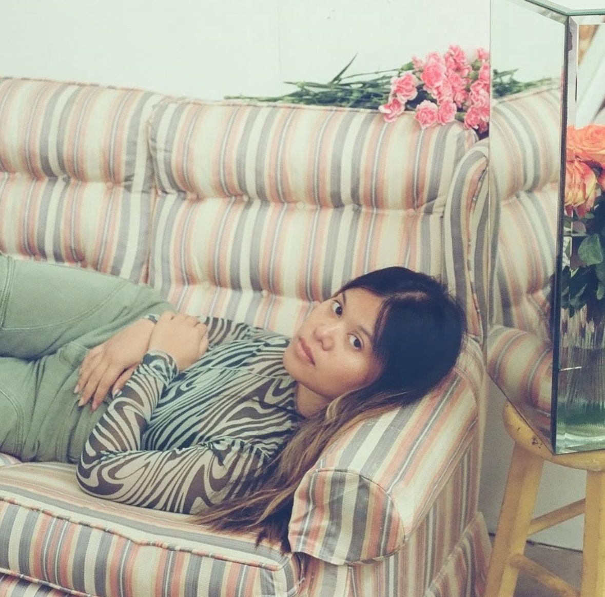 A person with brown hair lays on a pink and blue striped couch. They are wearing a black and white patterned shirt and green pants. 