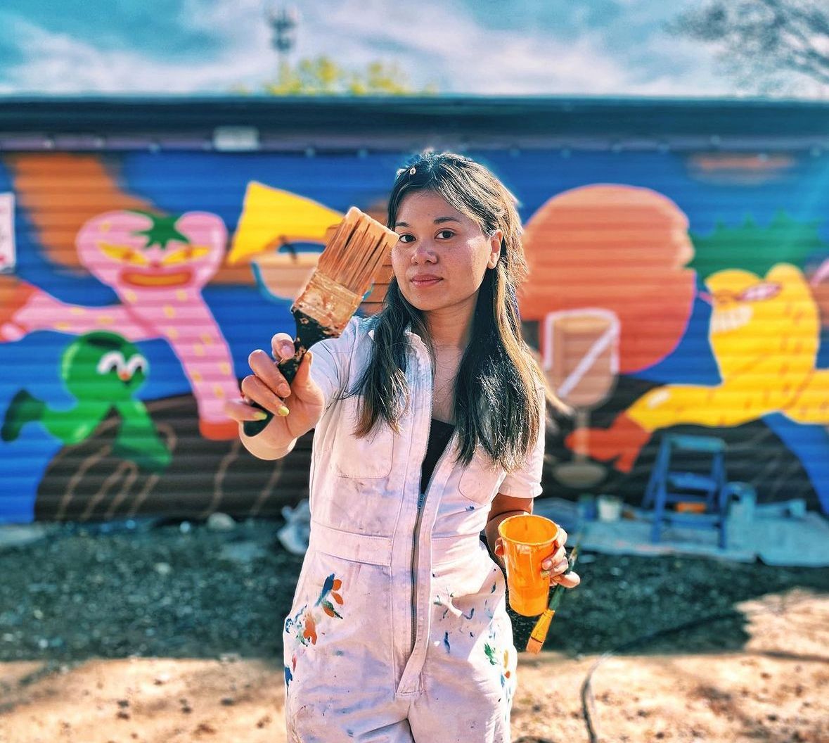 A person wearing white painter's overalls holds a brush with a mural they've completed behind them.