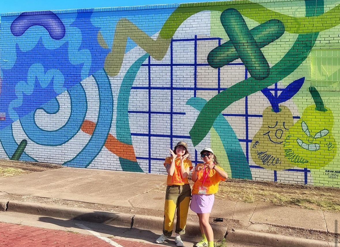 Two people stand in front of a mural with green and blue designs! 