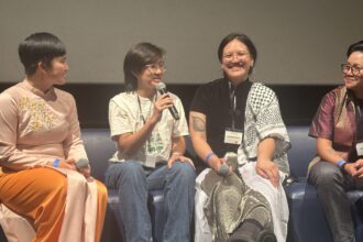 A panel of four individuals sit on a stage, with one person speaking into a microphone