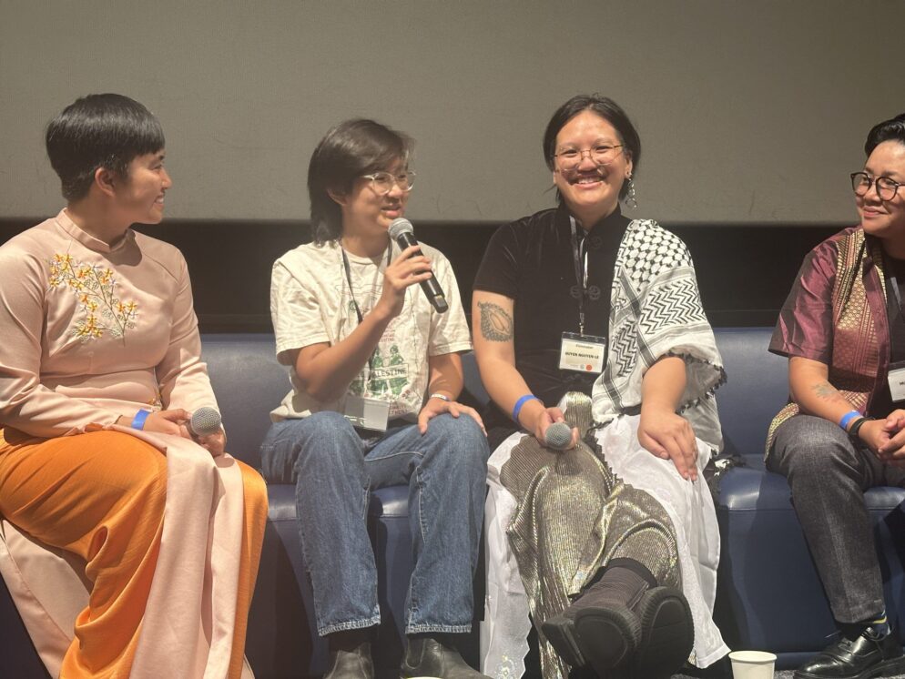 A panel of four individuals sit on a stage, with one person speaking into a microphone