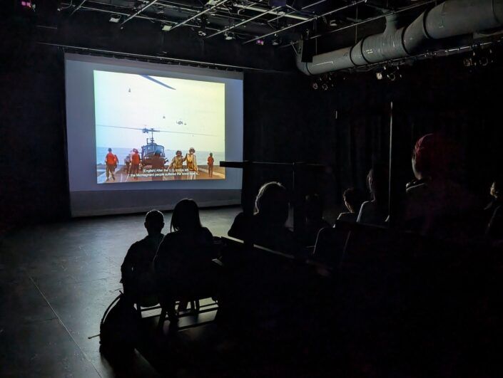 An audience in a darkened room looks at a screen depicting helicopters and soldiers