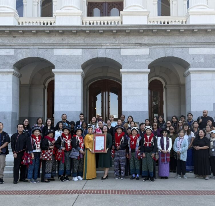 A large intergenerational crowd stands in front of a building, person is holding a plaque