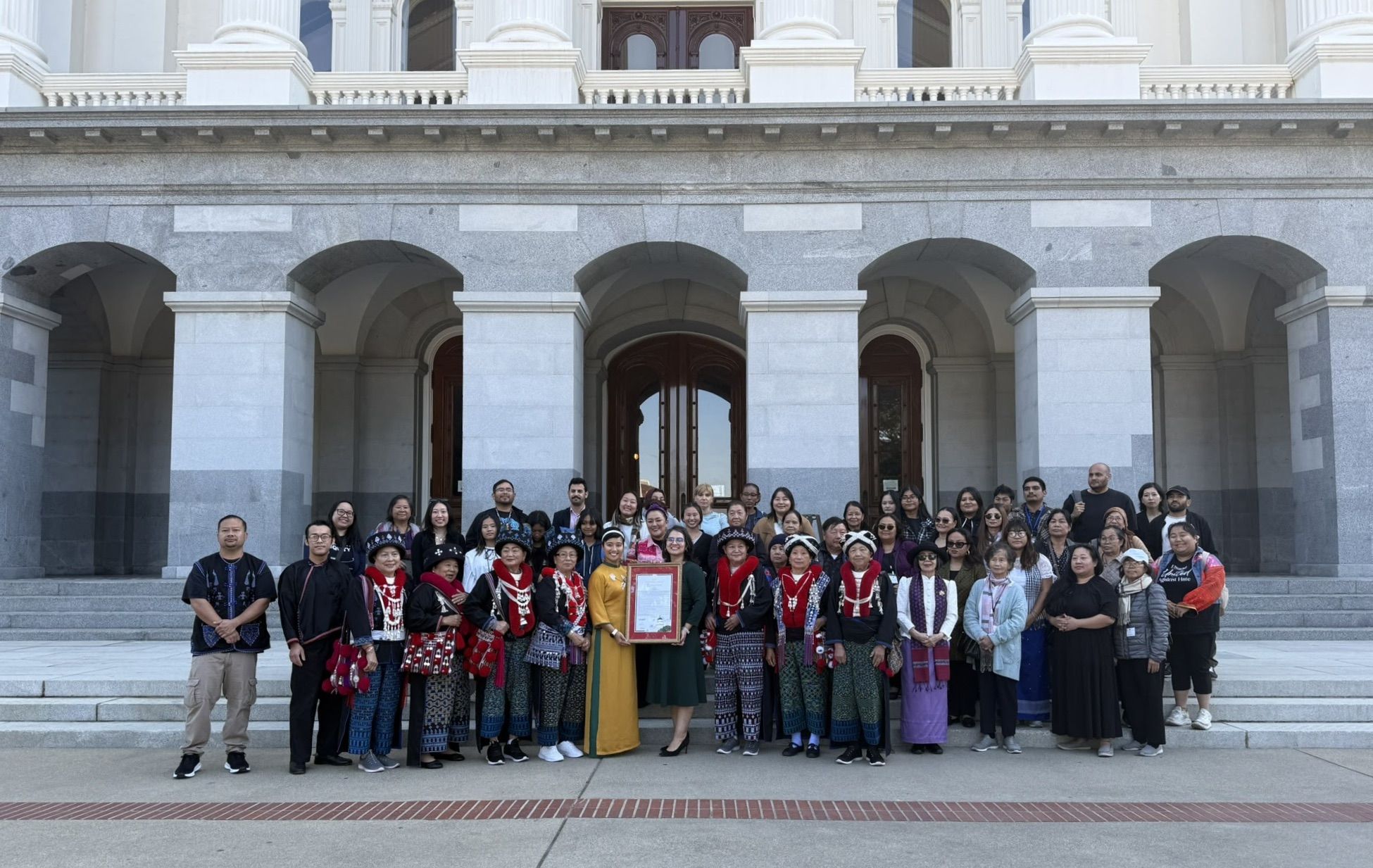 A large intergenerational crowd stands in front of a building, person is holding a plaque