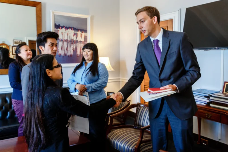 person shakes the hand of an individual wearing a suit and tie holding a folder and notebook