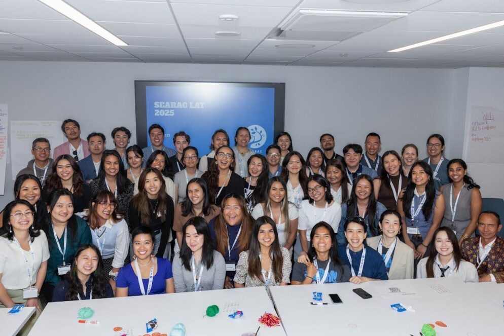 large group of individuals pose behind a gray table and in front of a blue screen